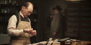 Movie still from “The Outfit” (2022), directed by Graham Moore – An older man in an apron working in a workshop; Medium shot, Over the shoulder angle