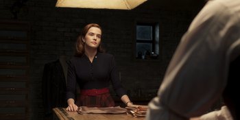 Movie still from “The Outfit” (2022), directed by Graham Moore – A woman sitting at a table in a dark room; Medium shot, Over the shoulder angle