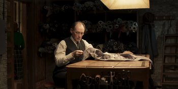 Movie still from “The Outfit” (2022), directed by Graham Moore – A man sitting at a table in front of rolls of fabric; Medium shot, Over the shoulder angle