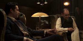 Movie still from “The Outfit” (2022), directed by Graham Moore – Two people sitting in a room with a lamp on the side of the room; Medium shot, Over the shoulder angle