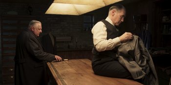 Movie still from “The Outfit” (2022), directed by Graham Moore – A man sitting on a table in a room; Medium shot, Over the shoulder angle