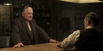 Movie still from “The Outfit” (2022), directed by Graham Moore – An older man sitting at a table in a room; Medium shot, Over the shoulder angle