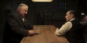 Movie still from “The Outfit” (2022), directed by Graham Moore – Two men sitting at a wooden table in front of a brick wall; Medium shot, Over the shoulder angle