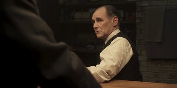 Movie still from “The Outfit” (2022), directed by Graham Moore – An older man sitting at a table in a room; Medium shot, Over the shoulder angle