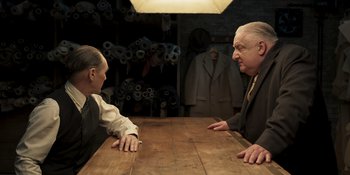 Movie still from “The Outfit” (2022), directed by Graham Moore – Two men sitting at a wooden table in front of each other; Medium shot, Over the shoulder angle