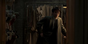 Movie still from “The Outfit” (2022), directed by Graham Moore – A man wearing a hat and a coat in a room full of clothes; Medium shot, High angle