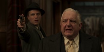 Movie still from “The Outfit” (2022), directed by Graham Moore – A man in a suit and tie holding a gun in front of another man in a suit and tie; Close Up shot, Over the shoulder angle