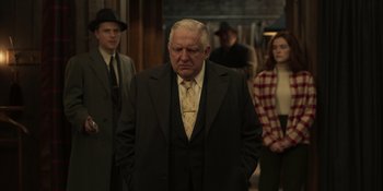 Movie still from “The Outfit” (2022), directed by Graham Moore – An older man in a suit and tie standing in front of a group of people; Medium shot, Over the shoulder angle
