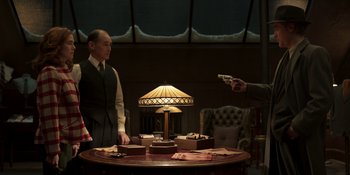Movie still from “The Outfit” (2022), directed by Graham Moore – A man holding a gun pointed at another man in front of a table; Medium shot, Over the shoulder angle