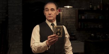 Movie still from “The Outfit” (2022), directed by Graham Moore – A man in a vest and tie holding a camera; Medium shot, High angle