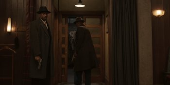 Movie still from “The Outfit” (2022), directed by Graham Moore – A man in a hat and a long coat standing in front of a door; Wide shot, Over the shoulder angle