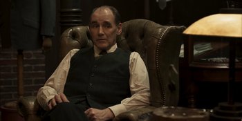 Movie still from “The Outfit” (2022), directed by Graham Moore – An older man sitting in a chair in a room; Medium shot, Over the shoulder angle