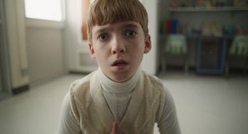 Movie still from “The Passenger” (2023), directed by Carter Smith – A young boy is sitting in a room with his hands in his chest; Close Up shot, Low angle