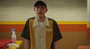 Movie still from “The Passenger” (2023), directed by Carter Smith – A young man wearing a hat and a brown and yellow shirt; Medium shot, Over the shoulder angle