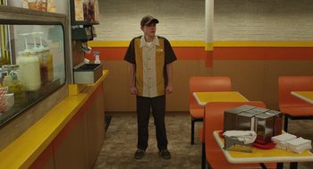 Movie still from “The Passenger” (2023), directed by Carter Smith – A man standing in front of a table in a fast food restaurant; Wide shot, High angle