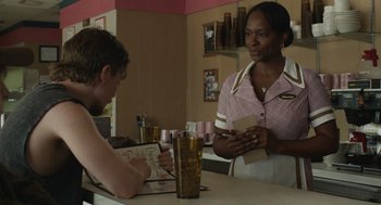 Movie still from “The Passenger” (2023), directed by Carter Smith – A woman standing at a counter talking to a customer; Medium shot, Over the shoulder angle