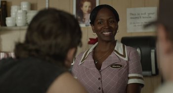 Movie still from “The Passenger” (2023), directed by Carter Smith – A woman in a pink and white dress smiling at a person; Close Up shot, Over the shoulder angle