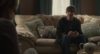 Movie still from “The Passenger” (2023), directed by Carter Smith – A man sitting on top of a couch holding a cell phone; Medium shot, Over the shoulder angle