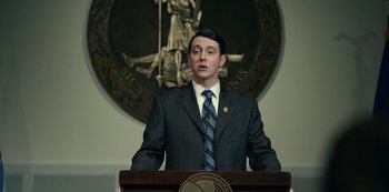 Movie still from “Dopesick” (2021), directed by Danny Strong – A man standing at a podium in front of a microphone; Medium shot, Low angle