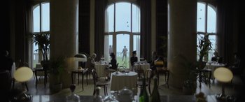 Movie still from “The Pigeon Tunnel” (2023), directed by Errol Morris – A group of people sitting at tables in front of a large window; Wide shot, Over the shoulder angle