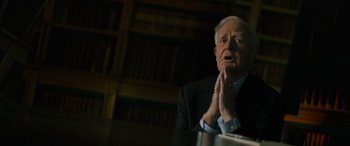 Movie still from “The Pigeon Tunnel” (2023), directed by Errol Morris – An older man in a black suit is sitting in front of a book shelf; Medium shot, Low angle