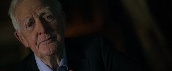 Movie still from “The Pigeon Tunnel” (2023), directed by Errol Morris – An older man wearing a suit and tie in the dark; Close Up shot, Over the shoulder angle