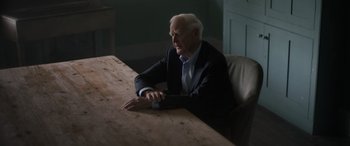 Movie still from “The Pigeon Tunnel” (2023), directed by Errol Morris – An older man sitting at a wooden table in a room; Medium shot, Over the shoulder angle