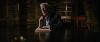 Movie still from “The Pigeon Tunnel” (2023), directed by Errol Morris – An older man sitting at a table in front of a book case; Medium shot, Over the shoulder angle
