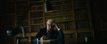 Movie still from “The Pigeon Tunnel” (2023), directed by Errol Morris – An older man is writing in a book in front of a bookshelf; Medium shot, High angle