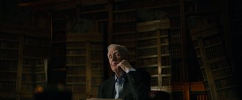 Movie still from “The Pigeon Tunnel” (2023), directed by Errol Morris – An older man sitting in a chair in front of a bookshelf; Medium shot, Low angle