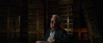 Movie still from “The Pigeon Tunnel” (2023), directed by Errol Morris – An older man sitting in front of a book shelf; Medium shot, Low angle
