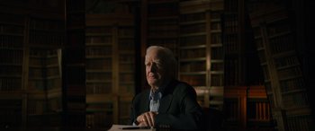 Movie still from “The Pigeon Tunnel” (2023), directed by Errol Morris – An older man sitting at a table in a library; Medium shot, Low angle
