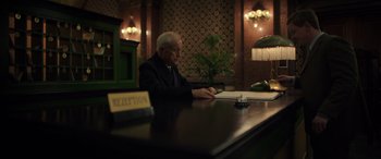 Movie still from “The Pigeon Tunnel” (2023), directed by Errol Morris – An older man sitting at a table in front of papers; Medium shot, Low angle