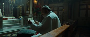 Movie still from “The Pope's Exorcist” (2023), directed by Julius Avery – An older man sitting in a church reading a book; Medium shot, Low angle