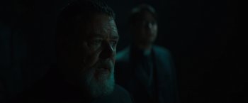Movie still from “The Pope's Exorcist” (2023), directed by Julius Avery – A man with a goatee and a beard is looking at the camera while another man stands in the background; Close Up shot, Over the shoulder angle