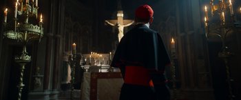 Movie still from “The Pope's Exorcist” (2023), directed by Julius Avery – A man in a priest's outfit standing in front of a crucifixion; Extreme Wide shot, Low angle