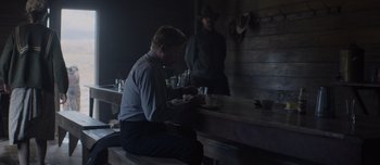 Movie still from “The Power of the Dog” (2021), directed by Jane Campion – A man sitting at a wooden table with a drink; Medium shot, Over the shoulder angle