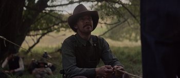Movie still from “The Power of the Dog” (2021), directed by Jane Campion – A man sitting in the grass wearing a cowboy hat; Medium shot, Over the shoulder angle