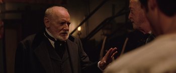 Movie still from “The Prestige” (2006), directed by Christopher Nolan – An older man wearing a suit and tie talking; Close Up shot, Over the shoulder angle