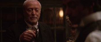 Movie still from “The Prestige” (2006), directed by Christopher Nolan – An older man holding a wine glass in a dimly - lit room; Close Up shot, Over the shoulder angle