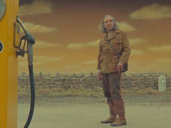 Movie still from “The Rat Catcher” (2023), directed by Wes Anderson – A man standing in front of a gas pump; Medium shot, Low angle