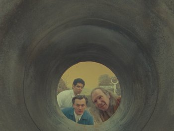 Movie still from “The Rat Catcher” (2023), directed by Wes Anderson – Three men are looking through a hole in a pipe; Medium shot, Low angle
