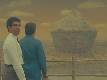 Movie still from “The Rat Catcher” (2023), directed by Wes Anderson – Two men are standing in front of a straw hut; Wide shot, Low angle