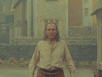Movie still from “The Rat Catcher” (2023), directed by Wes Anderson – An old man standing in front of a stone building; Medium shot, Low angle
