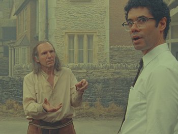 Movie still from “The Rat Catcher” (2023), directed by Wes Anderson – Two men standing next to each other in front of a stone building; Medium shot, Over the shoulder angle