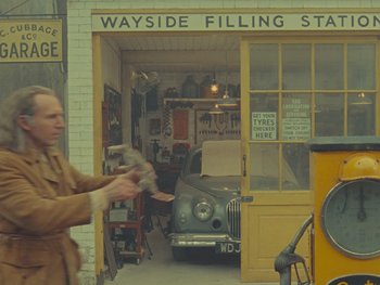 Movie still from “The Rat Catcher” (2023), directed by Wes Anderson – A man standing in front of a building with a car parked in front of it; Wide shot, High angle