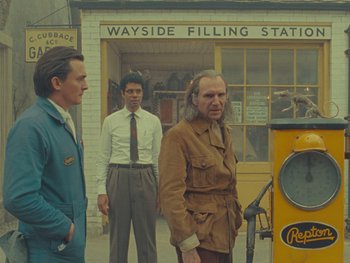 Movie still from “The Rat Catcher” (2023), directed by Wes Anderson – Three men standing in front of a gas pump; Medium shot, Low angle