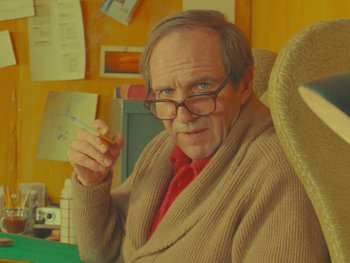 Movie still from “The Rat Catcher” (2023), directed by Wes Anderson – An older man sitting in a chair holding a cigarette in his hand; Medium shot, Over the shoulder angle