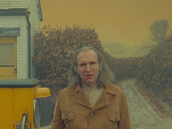 Movie still from “The Rat Catcher” (2023), directed by Wes Anderson – A man with long hair standing in front of a painting; Medium shot, Low angle