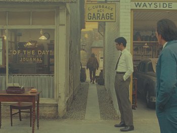 Movie still from “The Rat Catcher” (2023), directed by Wes Anderson – A man standing on the sidewalk in front of a building; Wide shot, Over the shoulder angle